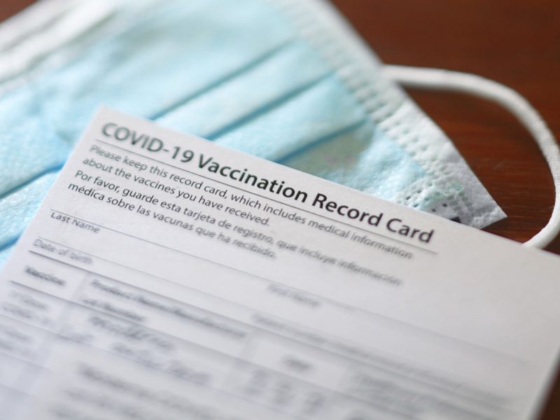A blank COVID-19 vaccination record card rests on top of a protective face mask. Photographed with a very shallow depth of field.